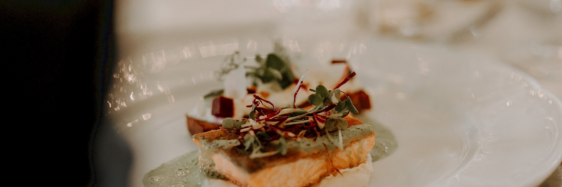 Schlossrestaurant Hellbrunn (c) Gassner Gastronomie, Fotografie Farbenspiel:  Auf dem Bild ist ein elegant angerichtetes Gourmetgericht zu sehen. Im Vordergrund liegt ein perfekt gegartes Lachsfilet auf einem Bett aus cremigem Püree, überzogen mit einer feinen grünen Soße und garniert mit frischen Microgreens. Dahinter befinden sich weitere Komponenten, möglicherweise geröstete Rote-Bete-Würfel und ein mit Kräutern dekorierter Frischkäse oder eine ähnliche Beilage. Das Ganze ist auf einem edlen Porzellanteller angerichtet und von elegantem Besteck umgeben – ein Gericht, das nicht nur geschmacklich, sondern auch visuell überzeugt. Ideal für ein festliches Dinner.