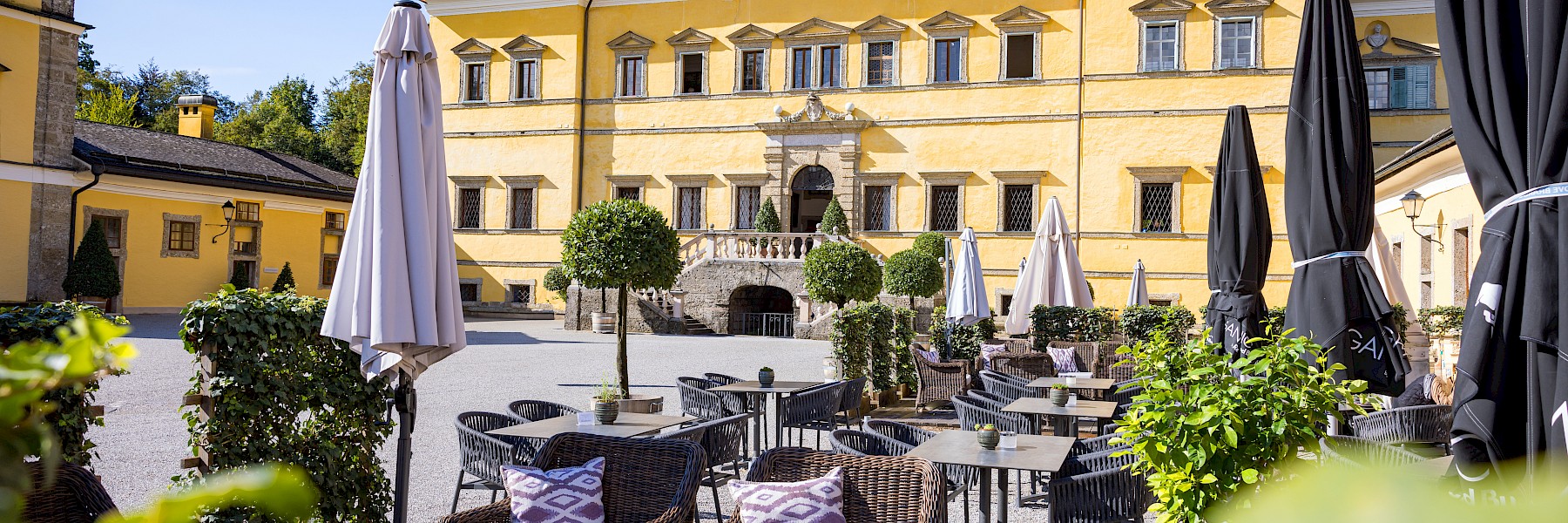 Schlosspark Hellbrunn (c) Gassner Gastronomie:  Das Bild zeigt eine sonnige Terrasse vor einem historischen, gelb gestrichenen Schlossgebäude mit barocker Architektur. Die stilvoll eingerichtete Außengastronomie ist mit gemütlichen Rattanmöbeln, violett-gemusterten Kissen und kleinen Topfpflanzen dekoriert. Große geschlossene Sonnenschirme stehen zwischen den Sitzgruppen, die durch niedrige Hecken voneinander getrennt sind. Im Hintergrund erhebt sich das prachtvolle Hauptgebäude mit symmetrisch angeordneten Fenstern, einer Steintreppe und gepflegten Buchsbaumkugeln. Der blaue Himmel und das helle Sonnenlicht verleihen der Szene eine freundliche und einladende Atmosphäre – ideal für eine entspannte Pause in historischer Umgebung.