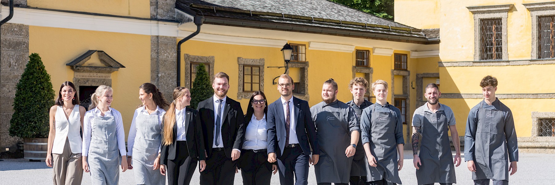 Das Bild zeigt das motivierte Team des Gasthauses zu Schloss Hellbrunn beim gemeinsamen Spaziergang im sonnendurchfluteten Innenhof. In harmonischer Mischung aus Servicekräften, Köchen und Management spiegeln ihre Outfits die Vielfalt der Aufgabenbereiche wider – von eleganten Anzügen bis hin zu professionellen Kochjacken. Die freundlichen Gesichter, das Lachen und die entspannte Haltung vermitteln Zusammenhalt, Freude an der Arbeit und echte Gastfreundschaft. Im Hintergrund sorgt die historische gelbe Schlossfassade für eine beeindruckende Kulisse, die Tradition und Atmosphäre vereint. Dieses Bild steht sinnbildlich für Teamgeist, Qualität und Herzlichkeit am Arbeitsplatz.