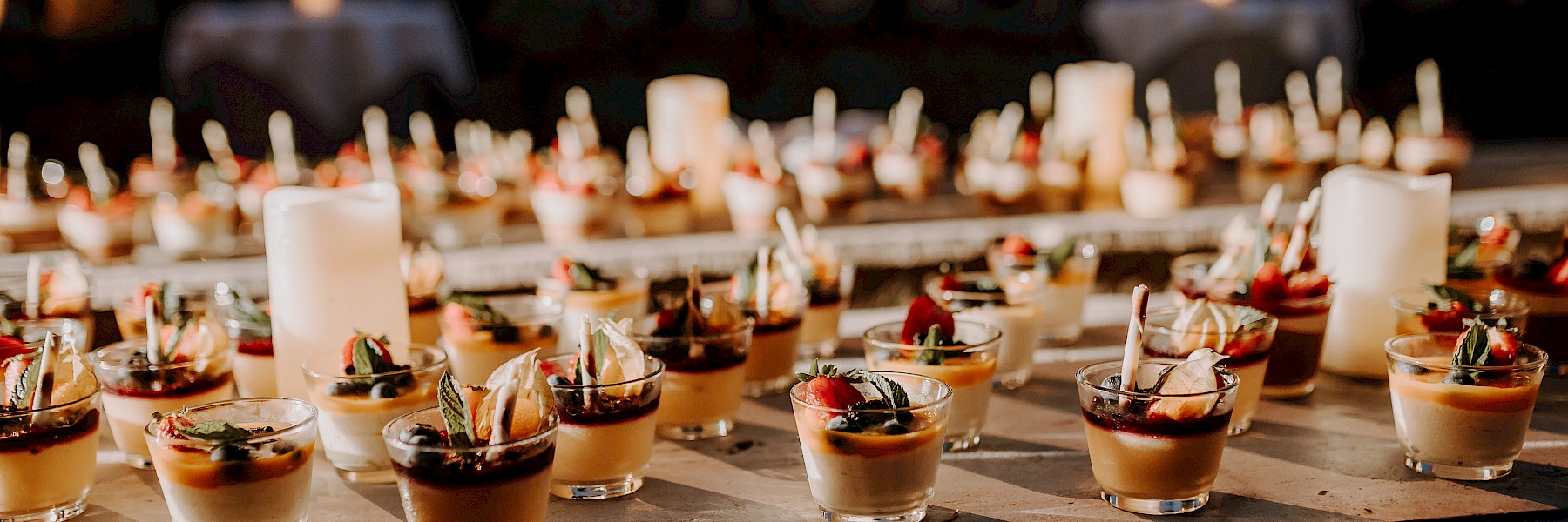 Dessertbuffet Hellbrunn (c) Gassner Gastronomie, Fotografie Farbenspiel: Das Bild zeigt eine festlich gedeckte Dessertstation bei Abendlicht. Zahlreiche kleine Gläser mit kunstvoll angerichteten Desserts – garniert mit Beeren, Minze, Sahne und weiteren feinen Zutaten – stehen ordentlich aufgereiht auf einem Tisch. Die sanfte, warme Beleuchtung schafft eine elegante und einladende Atmosphäre. Zwischen den Gläsern sorgen weiße Kerzen für zusätzliches Ambiente und stimmungsvolle Akzente. Die Szene wirkt wie aus einem hochwertigen Event oder einer Hochzeit und strahlt Liebe zum Detail und kulinarische Raffinesse aus. Ein Fest für die Augen und den Gaumen zugleich.