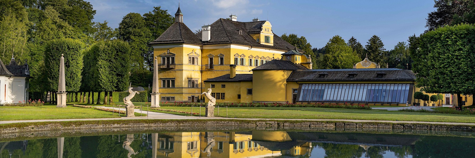 Hellbrunn Schloss Wasserparterre (c) Schlossverwaltung Hellbrunn Foto Auer: Das Bild zeigt Schloss Hellbrunn in Salzburg an einem sonnigen Tag, malerisch eingebettet in eine gepflegte Parklandschaft. Die barocke Architektur des gelben Schlosses spiegelt sich klar im ruhigen Wasser des davorliegenden Teichs. Umgeben von üppigem Grün, kunstvoll geschnittenen Hecken und eleganten Skulpturen, vermittelt das Ensemble eine friedliche und majestätische Atmosphäre. Besonders hervorstechend sind die dekorativen Fassadenelemente und das markante Dach mit kleinen Türmchen. Diese Szene strahlt historische Eleganz und zeitlose Schönheit aus – ein Ort, der Geschichte, Kunst und Natur harmonisch vereint.