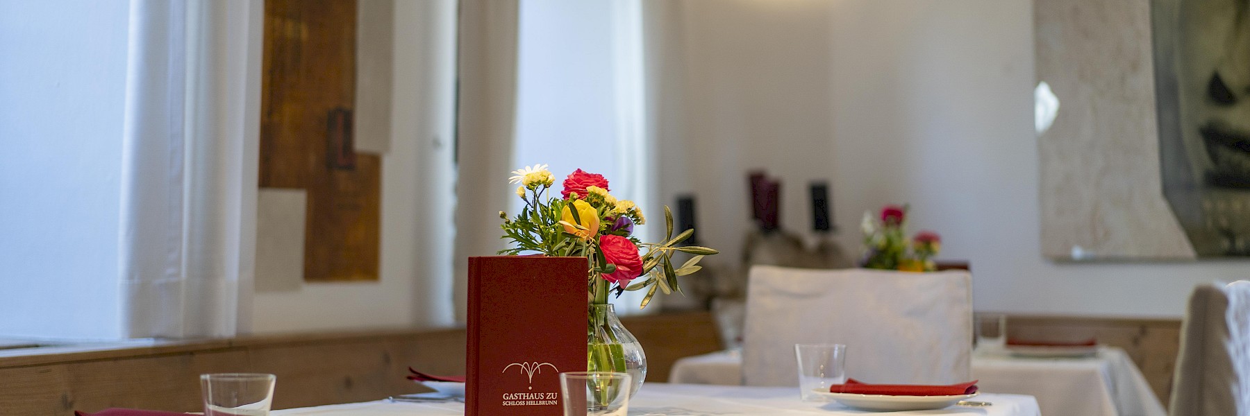 Gasthaus Hellbrunn (c) Gassner Gastronomie: Das Bild zeigt einen stilvoll gedeckten Innenraum des Gasthauses zu Schloss Hellbrunn. Die Tische sind mit weißen Tischdecken, roten Servietten und schlichten Gläsern eingedeckt. In der Mitte des Tisches steht ein Blumenstrauß in einer Glasvase, daneben eine dunkelrote Speisekarte mit dem Logo des Gasthauses. Der Raum wirkt hell und einladend, mit großen Fenstern und weißen Vorhängen. An der Wand hängen moderne Kunstwerke sowie ein Geweih, das für ein charmantes Zusammenspiel aus Tradition und zeitgenössischem Stil sorgt. Eine ruhige und elegante Atmosphäre prägt das Ambiente.