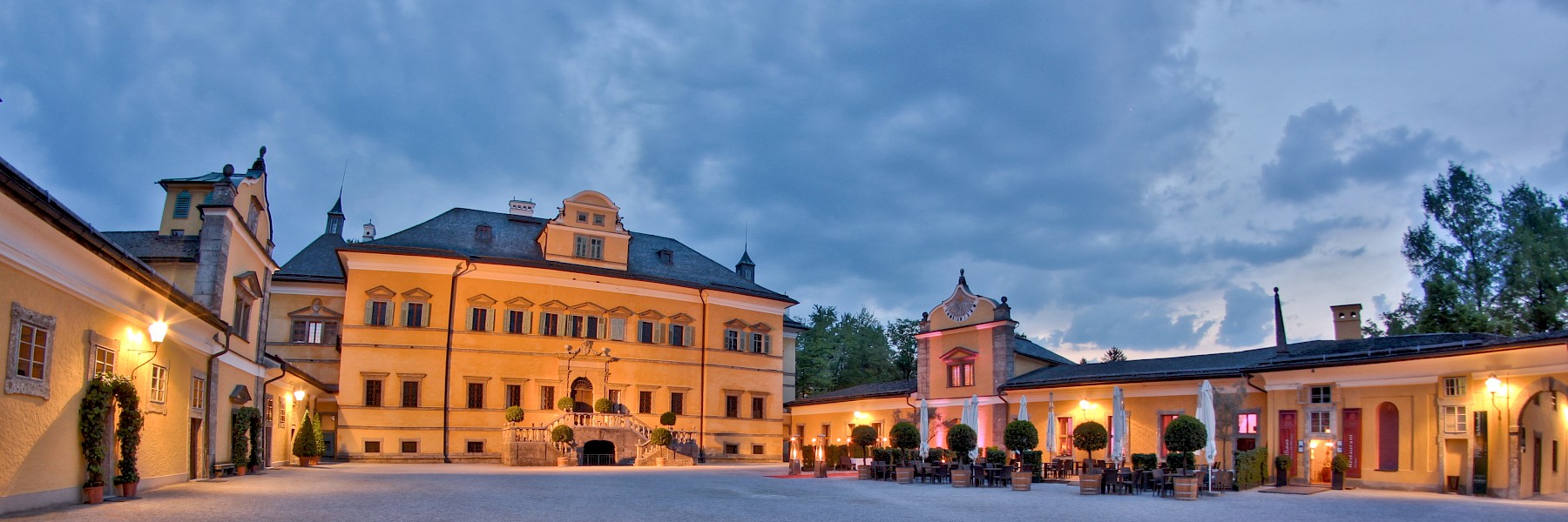 Eventlocation Hellbrunn: Das Bild zeigt den weitläufigen Innenhof von Schloss Hellbrunn in Salzburg in der Abenddämmerung. Das zentrale Schlossgebäude beeindruckt mit seiner symmetrischen Fassade, den kunstvoll verzierten Fenstern und der eleganten Freitreppe. Rechts im Bild befindet sich das festlich beleuchtete Gasthaus mit gemütlicher Außenterrasse, umrahmt von beleuchteten Bäumen und Heizstrahlern. Die warmen Gelbtöne der Gebäude kontrastieren stimmungsvoll mit dem blauen Abendhimmel. Der Platz wirkt ruhig und einladend – ein perfekter Ort für kulturelle Veranstaltungen, exklusive Dinners oder romantische Abende in historischem Ambiente.
