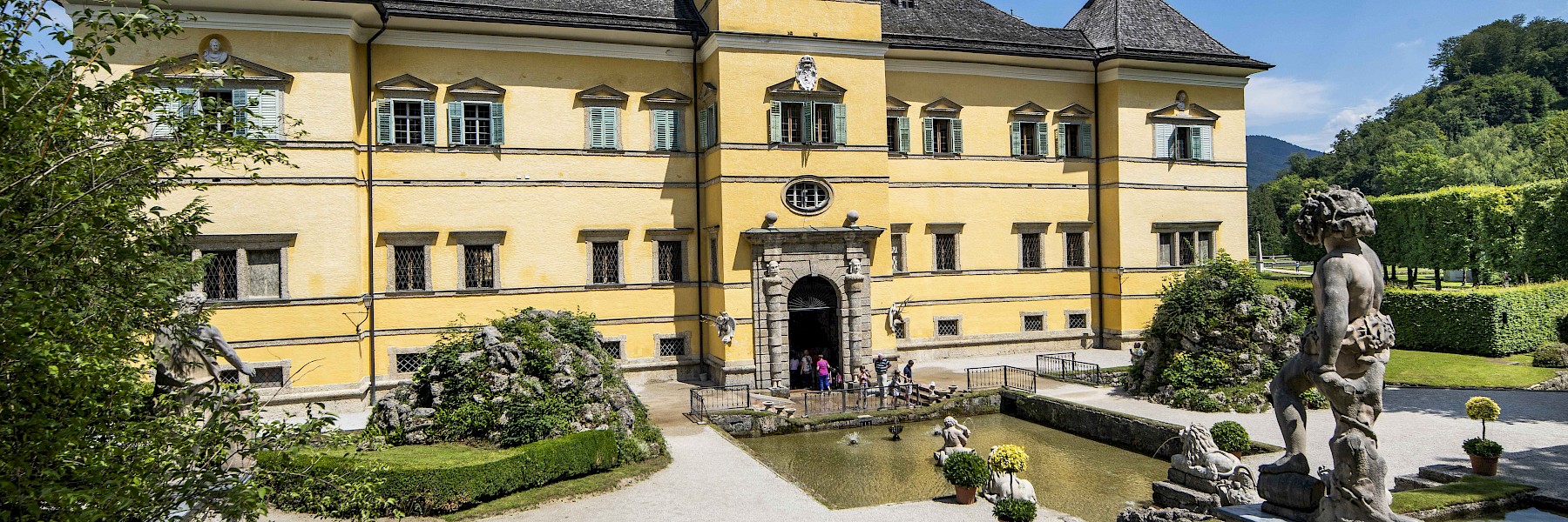 Hellbrunn Schloss (c) Schlossverwaltung Hellbrunn Foto Auer:  Das Bild zeigt die prachtvolle Fassade von Schloss Hellbrunn in Salzburg, einem barocken Lustschloss mit gelbem Anstrich und grünen Fensterläden. Vor dem Hauptportal erstreckt sich ein gepflegter Vorplatz mit einem rechteckigen Wasserbecken, gesäumt von Steinfiguren und symmetrisch angelegten Pflanzen. Besuchergruppen versammeln sich vor dem Eingang, während rundherum dichte Hecken, gepflegte Rasenflächen und kunstvolle Gartenelemente das historische Ambiente unterstreichen. Im Hintergrund erheben sich bewaldete Hügel unter einem klaren, blauen Himmel. Die Szene wirkt einladend und vermittelt den Charme und die Eleganz barocker Architektur inmitten idyllischer Natur.