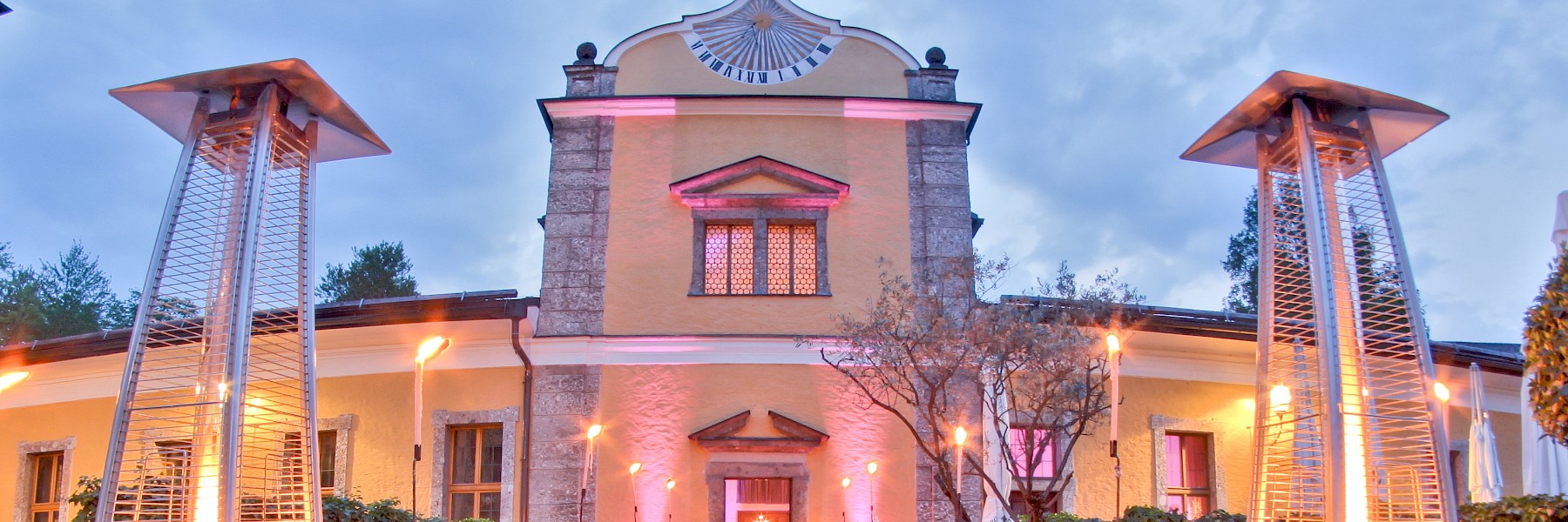 Gasthaus Hellbrunn (c) Gassner Gastronomie: Das Bild zeigt den festlich beleuchteten Eingangsbereich des Gasthauses zu Schloss Hellbrunn, geschmückt mit einem roten Teppich, der direkt zum Haupteingang führt. Beidseits säumen moderne Terrassenstrahler und Fackeln den Weg und erzeugen eine elegante, einladende Atmosphäre. Die Fassade erstrahlt in warmem Gelb und ist mit markanten Steinverzierungen geschmückt. Besonders ins Auge fällt der barocke Giebel mit einer kunstvollen Sonnenuhr. Vor dem Eingang stehen weiß gedeckte Tische – ein deutlicher Hinweis auf ein anstehendes Event oder ein stilvolles Abendessen.