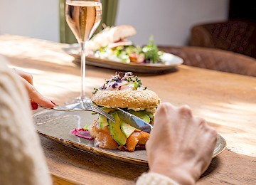 Das Bild zeigt eine köstliche Brunchkreation im Schlossrestaurant Hellbrunn. Im Fokus steht ein frischer Sesam-Bagel, liebevoll belegt mit Avocadoscheiben, zartem Räucherlachs, cremigem Frischkäse und einem pochierten Ei, garniert mit frischen Microgreens. Das Gericht wird auf einem stilvollen Keramikteller serviert, begleitet von einem Glas Sekt, was dem Moment eine festliche Note verleiht. Das warme Sonnenlicht und das rustikale Holzambiente schaffen eine einladende, entspannte Atmosphäre – perfekt für einen genussvollen Start in den Tag.