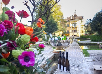 Das Bild zeigt eine elegante Outdoor-Location vor dem Schloss Hellbrunn, vorbereitet für einen stilvollen Empfang. Im Vordergrund stehen Flaschen mit Sekt oder Wein, zahlreiche Gläser und eine prunkvolle Schale mit Eis und gekühlten Getränken. Ein prachtvoller Blumenstrauß in lebendigen Farben ziert die Szene und sorgt für frühlingshaftes Flair. Die Kulisse mit dem barocken Schloss, dem Wasserbecken und den gepflegten Gärten im Hintergrund unterstreicht das edle Ambiente. Das warme Licht und die festliche Atmosphäre machen deutlich: Hier wird ein besonderes Ereignis stilvoll gefeiert.
