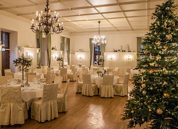 Der festlich geschmückte Saal strahlt in warmem Kerzenlicht und eleganter Zurückhaltung. Runde, weiß gedeckte Tische mit stilvollen Gedecken und winterlicher Floristik schaffen eine festliche Atmosphäre. Die cremefarbenen Stuhlhussen harmonieren perfekt mit der dezenten Wandgestaltung und den grünen Vorhängen. Im Raum verteilt stehen dekorative Kerzenleuchter, während ein großer, prachtvoll geschmückter Weihnachtsbaum mit goldenen Kugeln und Lichtern den rechten Bildrand ziert. Über allem hängen elegante Kronleuchter, die dem Raum einen Hauch von Noblesse verleihen. Der Saal ist ideal für ein stilvolles Weihnachtsdinner oder eine festliche Winterhochzeit.