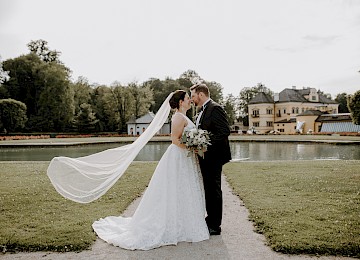 Das Bild zeigt ein romantisches Hochzeitspaar vor der eindrucksvollen Kulisse von Schloss Hellbrunn. Die Braut trägt ein elegantes, weißes Kleid mit fließendem Schleier, der sanft vom Wind erfasst wird, während der Bräutigam klassisch im schwarzen Anzug gekleidet ist. Beide stehen sich innig gegenüber, mit einem See und dem historischen Schloss im Hintergrund. Die grüne Parklandschaft, das Wasser und die ruhige Atmosphäre unterstreichen die besondere Magie dieses Moments – ein perfektes Motiv für eine stilvolle Hochzeitskulisse inmitten historischer Natur.