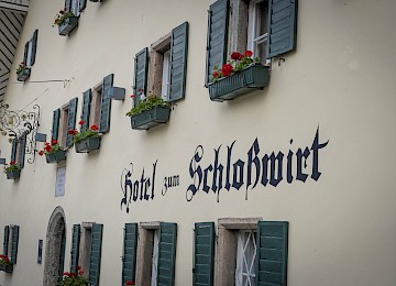 Das Bild zeigt die Fassade des traditionellen „Hotel zum Schlosswirt“ mit einem charmanten, historischen Flair. Die cremefarbene Wand ist mit grünen Fensterläden verziert, an denen üppige Blumenkästen mit roten Geranien hängen – ein typisches Bild alpenländischer Gastfreundschaft. Die dekorative Schmiedeeisenbeschilderung und die gotische Schriftart des Hotelnamens verstärken den nostalgischen Charakter. Das Gebäude vermittelt eine gemütliche, einladende Atmosphäre und wirkt gepflegt und liebevoll gestaltet – ideal für Reisende, die Authentizität und Ruhe in einem traditionsreichen Haus suchen.