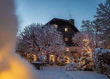 Das Bild zeigt eine stimmungsvolle Winterabend-Szene vor einem idyllischen Haus, das in weihnachtlichem Lichterglanz erstrahlt. Schneebedeckte Bäume, geschmückt mit roten Kugeln, und festlich beleuchtete Tannenbäume schaffen eine märchenhafte Atmosphäre. Warmes Licht dringt durch die Fenster und vermittelt Geborgenheit und Gemütlichkeit. Weiße Gartenmöbel im Schnee verstärken den Eindruck eines friedlichen, festlichen Rückzugsorts. Rauch steigt aus dem Kamin und rundet das romantische Winterbild ab – eine perfekte Kulisse für besinnliche Momente und zauberhafte Festtage inmitten einer verschneiten Landschaft.