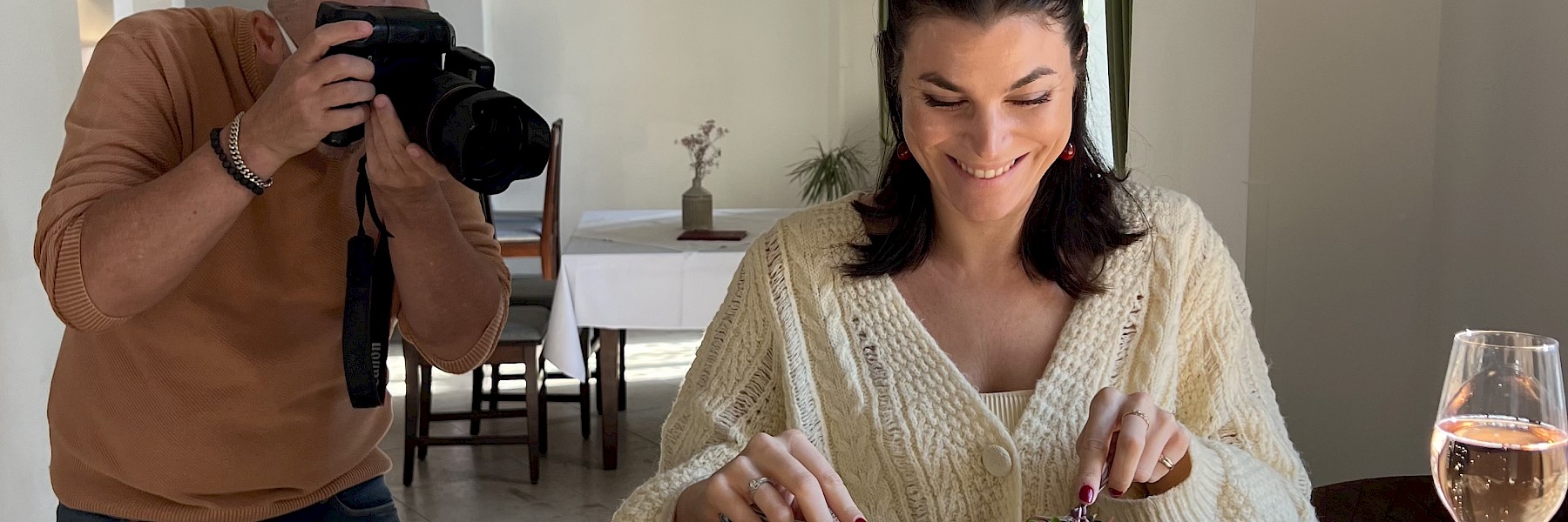 Hellbrunn Schloss Wasserparterre (c) Schlossverwaltung Hellbrunn Foto Auer: Das Bild zeigt eine junge Frau, die in einem stilvollen, lichtdurchfluteten Raum einen kunstvoll angerichteten Burger genießt. Sie lächelt, während sie das Gericht mit Messer und Gabel anschneidet. Rechts von ihr steht ein Glas Rosé-Sekt. Links im Bild hält ein Fotograf mit konzentriertem Blick eine professionelle Kamera und fängt den Moment ein. Die Szene wirkt natürlich und lebendig – eine Mischung aus kulinarischem Genuss und professioneller Food-Fotografie. Die harmonische Lichtstimmung und das elegante Ambiente unterstreichen die hochwertige Atmosphäre des Settings.