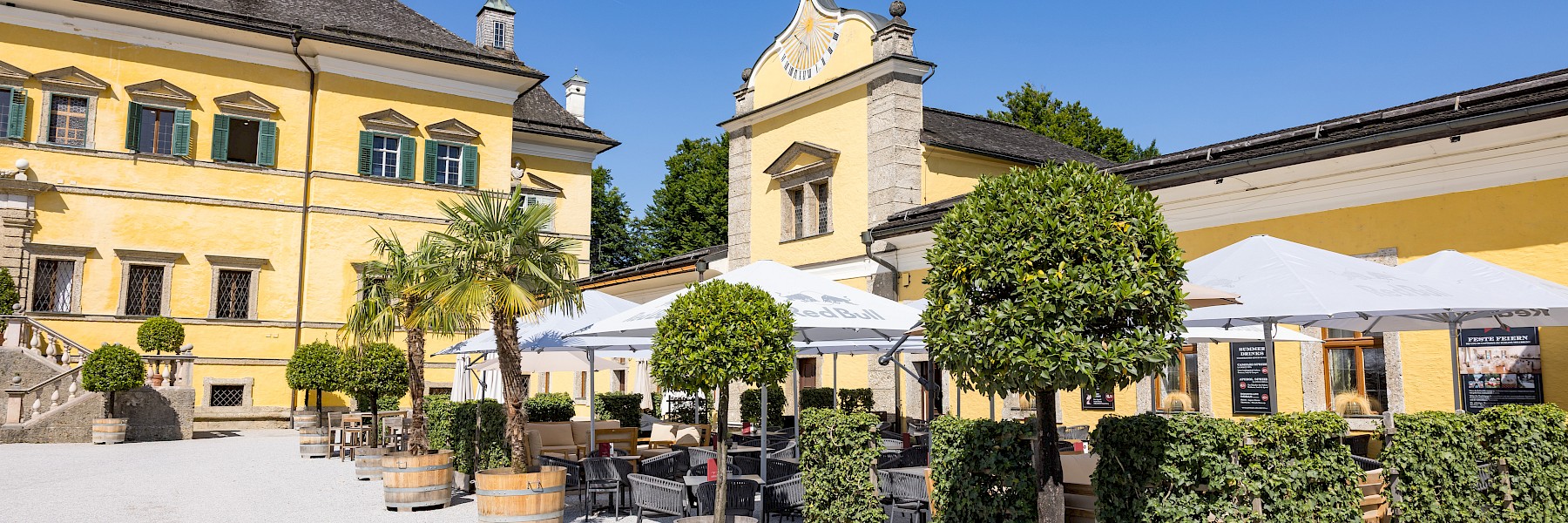 Hellbrunn Schloss (c) Schlossverwaltung Hellbrunn Foto Auer:  Das Bild zeigt die prachtvolle Fassade von Schloss Hellbrunn in Salzburg, einem barocken Lustschloss mit gelbem Anstrich und grünen Fensterläden. Vor dem Hauptportal erstreckt sich ein gepflegter Vorplatz mit einem rechteckigen Wasserbecken, gesäumt von Steinfiguren und symmetrisch angelegten Pflanzen. Besuchergruppen versammeln sich vor dem Eingang, während rundherum dichte Hecken, gepflegte Rasenflächen und kunstvolle Gartenelemente das historische Ambiente unterstreichen. Im Hintergrund erheben sich bewaldete Hügel unter einem klaren, blauen Himmel. Die Szene wirkt einladend und vermittelt den Charme und die Eleganz barocker Architektur inmitten idyllischer Natur.